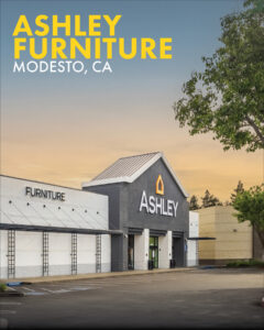 Exterior of the newly completed Ashley Furniture store in Modesto, CA at dusk, featuring a dark gray peaked entrance, a white brick facade, and yellow text overlay.